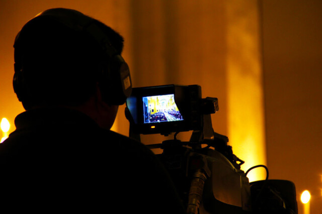 A person wearing headphones operates a video camera, capturing an event in a well-lit hall. The camera's viewfinder displays a scene with an audience and illuminated surroundings. Person with headphones operating video camera, filming event in illuminated hall.