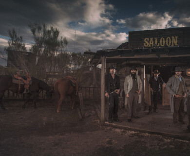 A group of men dressed in 19th-century Western attire stands in front of a wooden saloon. Two horses are tied nearby under a cloudy sky, adding to the rustic setting. Men in 19th-century Western attire by a saloon with horses under a cloudy sky.