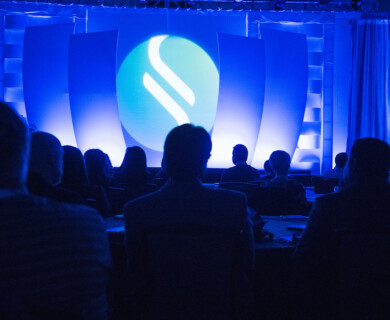 An audience is seated in a dimly lit room facing a stage with a large screen displaying a blue and white logo. The environment suggests a formal conference or presentation setting, using dramatic lighting effects. Audience in dim room facing a stage with a blue and white logo on a screen.