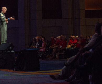 A person wearing a military flight suit is speaking on a stage in front of an audience seated in a dimly lit auditorium. The audience consists of individuals in casual and formal attire, some taking notes. The speaker appears to be engaged in a presentation or lecture. Person in flight suit speaking to audience in dim auditorium during presentation.