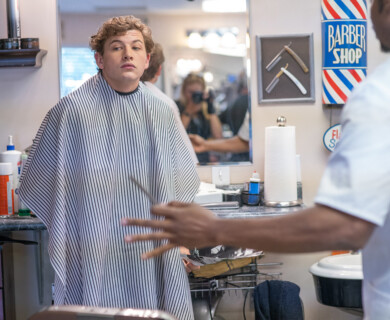 A person sits in a barber chair wearing a striped cape, looking towards the mirror. A barber, partially visible, is standing nearby, holding a tool. The setting features various barbering equipment and decor, including a "Barber Shop" sign on the wall. Person in a barber chair with a striped cape, barber holding tool, shop decor visible.