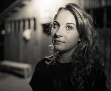 A black and white portrait of a person with long hair, standing outdoors at night. The background is softly blurred, showing a structure with vertical lines. The lighting highlights the person's facial features. Black and white portrait of a person with long hair outdoors at night, blurred vertical lines.