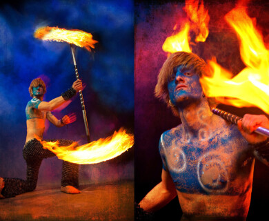 Performer painted blue with a flaming staff in a colorful, dramatic pose.