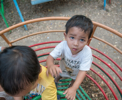 Three children are playing on a colorful climbing structure in an outdoor setting. One child is looking up at the camera, while the others are engaged in climbing. The ground is covered with dry leaves and dirt. Children playing on a colorful climbing structure outdoors with leaves on the ground.