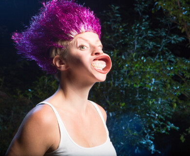 Person with spiky, colorful hair making a face in front of dark foliage outdoors.