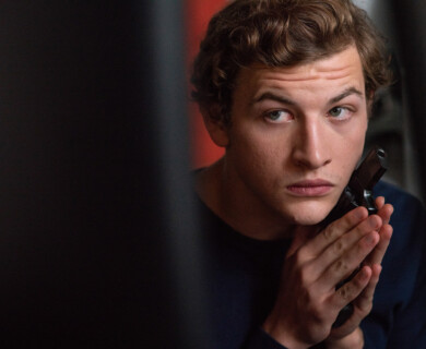 A person with curly hair holds a handgun close to their cheek, appearing thoughtful or pensive. The background is blurred, drawing focus to the individual's expression. Person with curly hair holds handgun near cheek, thoughtful expression, blurred background.