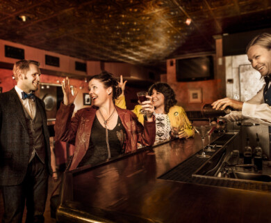 Four people in a dimly lit bar, bartender pouring drinks, patrons conversing cheerfully.