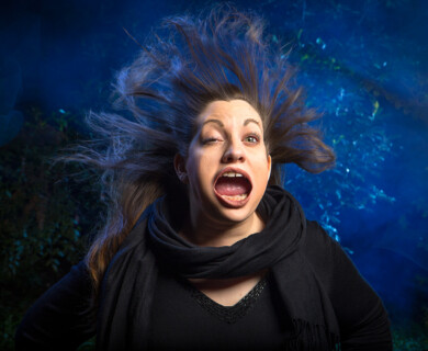 Person in expressive pose with open mouth, eyes wide, against dark, blue-green background.