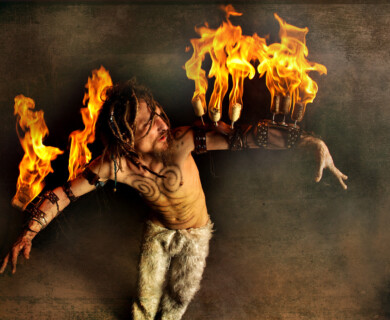 Performer in fur-like pants with arm-mounted fire torches, dreadlocks, and body paint.