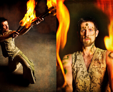 Man with body paint performs fire act, holding torches, wearing patterned clothing, fiery backdrop.