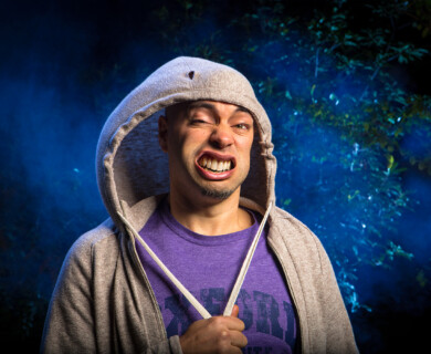Person in gray hoodie with funny expression, dark background with blue tint and faint foliage.