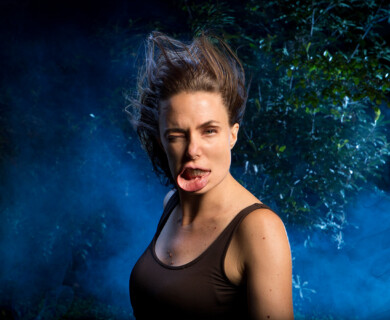 Person making a funny face with tongue out, one eye closed, against blue smoke and foliage.