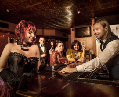 Group at a dimly lit bar, bartender serving drink to person in corset, lively atmosphere.