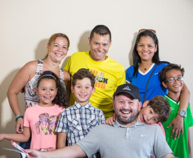 A group of eight people, including adults and children, is gathered for a cheerful photo indoors. They are standing closely together, wearing casual clothing, and smiling at the camera. Group of eight, indoors, adults and children smiling together in casual clothing