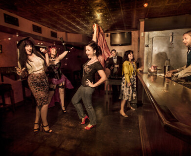 Lively bar scene with vibrant outfits, playful interactions, and a bartender engaging with guests.