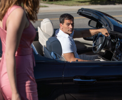 A man is seated in the driver's seat of a black convertible car, looking back towards a woman in a pink dress walking nearby. The car's interior is cream-colored, and the setting appears to be a daytime street scene. Man in black convertible looks back at woman in pink dress on street, cream car interior visible.