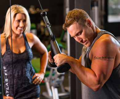 A man and a woman are inside a gym. The man is using a cable machine for exercise, focusing on his workout. The woman is standing nearby, watching him with a smile. The gym environment includes exercise equipment and weights. Man exercises with cable machine in gym, woman observes smiling.