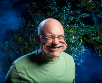 Man in green shirt outdoors with humorously distorted face, bluish foliage background.