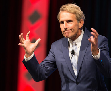 A man in a blue suit gestures expressively during a speech or presentation. He wears a light-colored tie, and a microphone is clipped to his lapel. The background features red patterns on a dark backdrop. Man in blue suit gestures during speech with red patterned backdrop and lapel microphone