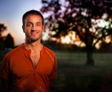 A man wearing an orange shirt is standing outdoors in a park setting during sunset. The background features a blurred silhouette of a tree with a sky displaying warm colors. Man in orange shirt stands in a park at sunset, blurred tree in the background.