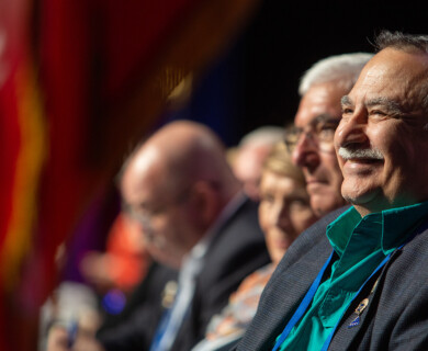 A man in a suit and teal shirt, seated in a row with other people, appears to be smiling at an event. The setting suggests a formal gathering with an audience. Man in suit and teal shirt smiling at a formal event with others seated nearby.