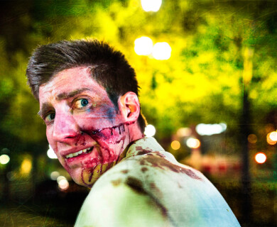 Man with theatrical cuts, bruises makeup, stained shirt, outdoors, blurred lights and greenery.