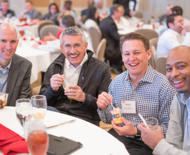 A group of four men are sitting together at a banquet table, enjoying desserts in a social or professional gathering. The setting appears to be a conference or event, with others present in the background. They are smiling and appear to be engaged in conversation. Four men at a banquet table enjoying desserts, smiling, and conversing at an event.
