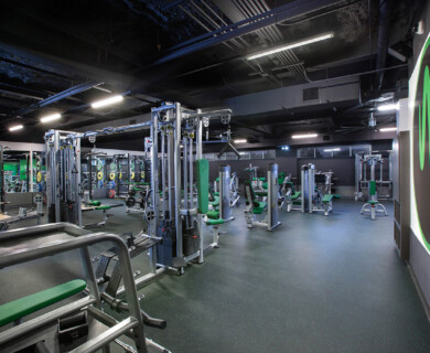 A modern gym features various exercise machines, including cable machines and weightlifting equipment, arranged in a spacious, well-lit environment with green accents on equipment and walls. Modern gym with well-lit exercise machines, including cable and weightlifting equipment.