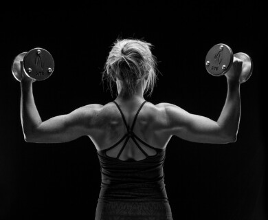 A black and white photo showing a person from behind, lifting a dumbbell in each hand. The person is wearing a tank top, highlighting muscular arms and back, set against a dark background. Person in tank top lifting dumbbells, showcasing muscular arms and back, dark background.