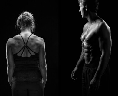 The image is a black and white composition featuring a woman and a man. The woman is seen from the back wearing a fitness top with straps, highlighting her muscular build. The man is shown shirtless, facing sideways, showcasing a well-defined muscular physique. Both individuals are set against a dark background. Man and woman in black and white, showcasing muscular physiques against dark background.