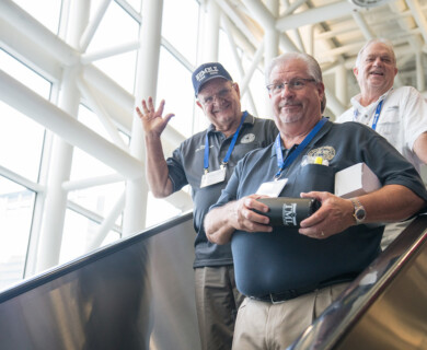 Three older men are standing on an escalator indoors, smiling and waving. Two are wearing dark polo shirts with identification badges, and one is holding a black tumbler. The setting appears to be a modern building with large windows and metal beams. Three older men on an escalator indoors, smiling and waving in a modern building.