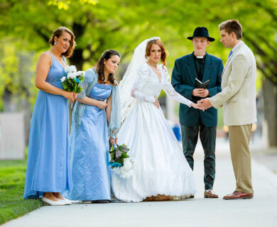 Outdoor wedding ceremony with bride, groom, minister, and bridesmaids on a tree-lined path.
