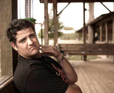 A person is leaning against a wooden post on a covered porch, looking towards the camera. There is a grassy area and an old building in the background. The person is wearing a black T-shirt and has a few bracelets on their wrist. Person in black T-shirt leaning against a wooden post on a porch, grassy area and building behind.
