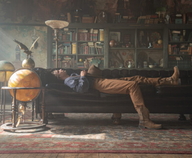 A person is lying on a black leather couch in a cozy, dimly lit room. The room features a large bookshelf filled with books and various decorative objects. Two globes are positioned near the couch, and sunlight streams through a window, creating a warm ambiance. Person lying on black leather couch in room with bookshelf, globes, and sunlight streaming in.