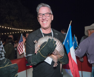 A person wearing gloves is smiling while holding an armadillo. They are at an outdoor event decorated with string lights and various flags, including the American flag. Other people are present in the background. Person with gloves holding an armadillo at event with string lights and American flag.