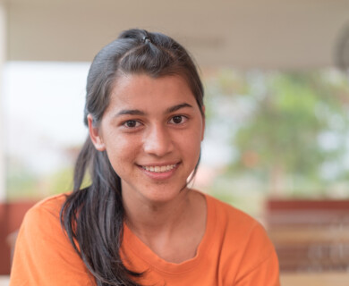A person with long dark hair wearing an orange shirt is smiling at the camera. The background is blurred, suggesting an outdoor setting with greenery. Person with long dark hair in an orange shirt smiling outdoors with a blurred green background.