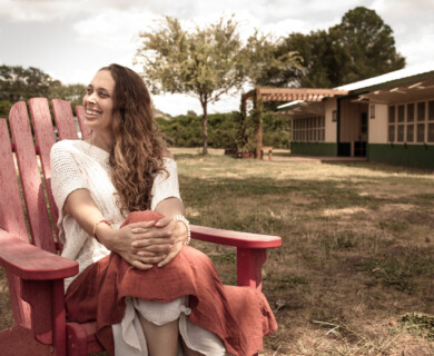 A person with long hair is sitting on a red wooden chair outdoors, smiling. They are wearing a white sweater and a burnt orange skirt. In the background, there is a green and white building and some trees under a partly cloudy sky. Person with long hair sits smiling on red chair outdoors, wearing a white sweater and orange skirt.