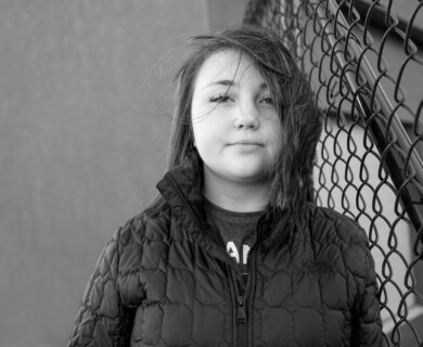 A black and white portrait of a person wearing a quilted jacket, standing next to a chain-link fence. The person's hair is slightly tousled. Person in a quilted jacket by a chain-link fence, hair slightly tousled, black and white portrait.