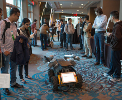 A group of people is gathered around a small robotic vehicle indoors. The rover-like machine is equipped with robotic arms and a display screen. The setting seems to be a conference or exhibition area, with attendees observing the robot's features. Group examining robotic rover with arms and screen at indoor conference or exhibition.