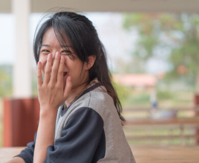 A person with dark hair is smiling while partially covering their face with one hand. They are sitting in an outdoor setting with blurred greenery in the background. Person with dark hair smiling, covering face with hand, blurred greenery background.