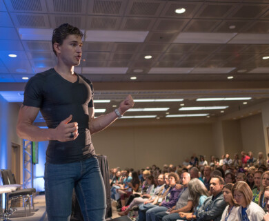 A person is speaking on stage in front of an audience inside a conference room. The speaker is wearing a microphone headset and gesturing with their hands. The audience is seated, attentively listening in a brightly lit room with rows of chairs and a ceiling with recessed lighting. Person speaking on stage with a headset in a conference room, audience listening attentively.