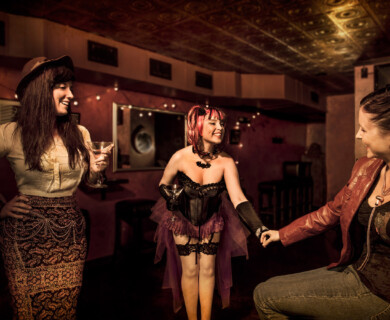 Three women in a dimly lit bar, all holding drinks, with string lights in the background.