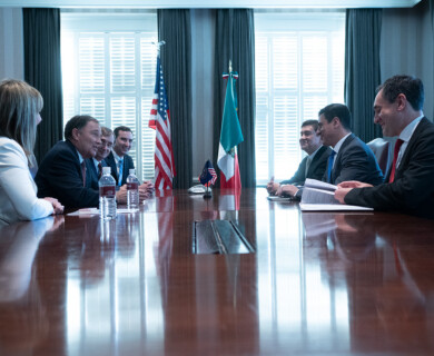 A group of people is sitting at a long conference table engaged in a meeting. The room features two national flags, the United States and Mexico. Some individuals have documents and water bottles on the table, and there are smiles and discussions taking place. The setting appears formal, with large windows in the background. Meeting at a conference table with US and Mexico flags, people discussing with documents and water.