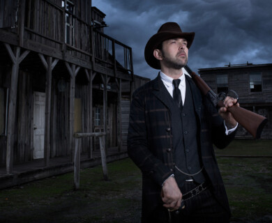 A person dressed in a vintage-style suit and hat stands outdoors holding a rifle. The background features wooden buildings with a rustic, western appearance, under a cloudy sky. Person in vintage suit holds rifle outside rustic wooden buildings under cloudy sky.