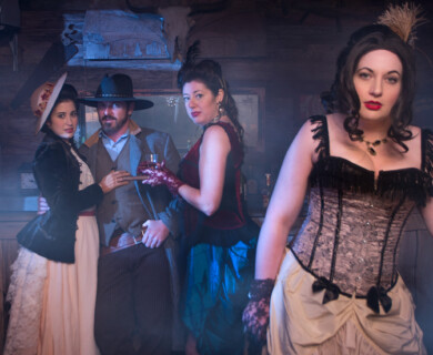 A group of individuals dressed in vintage Western-style attire are in a dimly lit, rustic saloon setting. One person wears a wide-brimmed hat, while others are in period dresses and corsets. The scene is atmospheric, with visible smoke or haze enhancing the historical theme. Group in vintage Western attire in a smoky saloon setting.