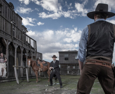 A scene depicting a classic Western standoff is set in a rustic town. Two cowboys in traditional attire face each other, their hands poised over their pistols. A horse is tethered nearby, and a woman dressed in period clothing stands on the wooden porch of a weathered building, observing the confrontation. The sky is partly cloudy, adding to the dramatic atmosphere. Western standoff in rustic town, two cowboys poised, woman observing, horse tethered nearby