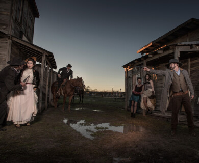 A dramatic scene set in a Western-style town depicts a tense confrontation. On the left, a man appears to be threatening a woman against the wall of a wooden building. In the center, a person on horseback enters the scene. To the right, a man is aiming a gun, while two women and another man stand nearby observing the situation. The setting suggests a storyline from a historical or cinematic Wild West theme. Western-style town scene with tense standoff; man on horseback, others in dramatic poses.
