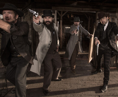 Four men dressed in Wild West-style attire, including hats and long coats, are standing on a wooden porch. They are holding pistols and appear to be aiming toward a target outside the image frame. The setting is reminiscent of a historical Western scene. Four men in Wild West attire with pistols aiming on a wooden porch, historical Western scene.