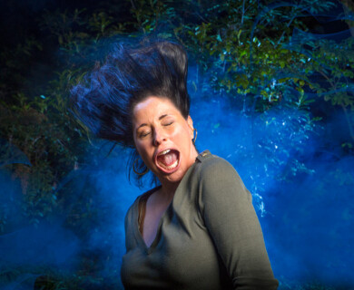 Woman with flowing hair and open mouth against blue mist and green foliage background.