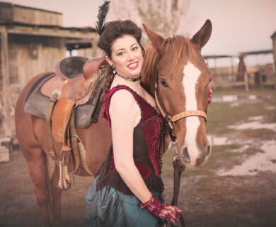 A woman dressed in period costume stands next to a brown horse with a white face marking. The setting appears to be a rustic outdoor location with wooden buildings in the background, evoking a historical or western theme. Woman in period costume beside horse with white marking, rustic wooden buildings in background.